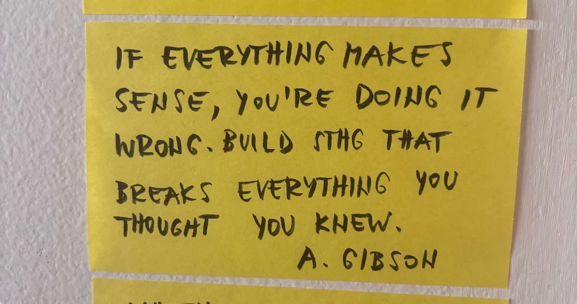 A photo of a yellow post-it mounted to a white wall. It reads: 'If everything makes sense, you're doing it wrong. Build something that breaks everything you thought you knew.' by Andrea Gibson